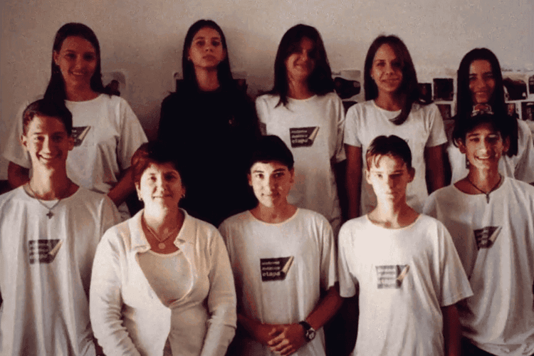 Foto de grupo em ambiente interno: nove pessoas posam para a câmera, cinco em pé ao fundo e quatro à frente, usando camisetas brancas com um logotipo; ao fundo há uma parede clara com algumas imagens fixadas.