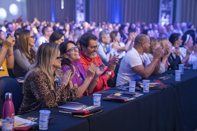 Plateia em um auditório durante um evento: pessoas sentadas em mesas com copos e materiais, aplaudindo e sorrindo sob iluminação azul.