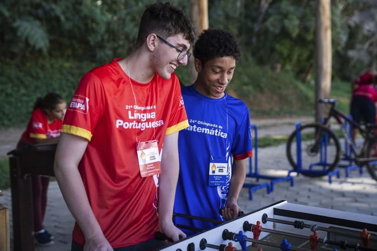 Dois jovens com crachás se divertem jogando pebolim ao ar livre; um veste camiseta vermelha da Olimpíada de Língua Portuguesa e o outro camiseta azul da Olimpíada de Matemática, com área verde ao fundo.