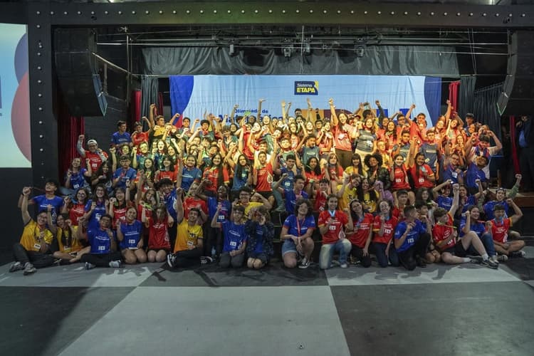 Grande foto de grupo em um palco: dezenas de estudantes com camisetas coloridas e crachás levantam os braços e comemoram, com um painel do Sistema Etapa ao fundo.