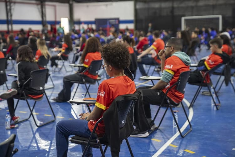Alunos sentados em cadeiras individuais em um ginásio, usando camisetas vermelhas do Sistema Etapa, durante as Olimpíadas do Sistema Etapa; ao fundo há várias fileiras de participantes.