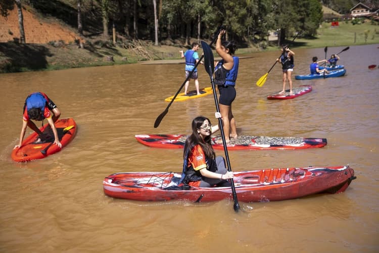Jovens praticam atividades aquáticas em um lago: uma pessoa rema em um caiaque vermelho em primeiro plano, enquanto outras usam pranchas de stand up paddle e um caiaque ao fundo, com árvores e área verde ao redor.