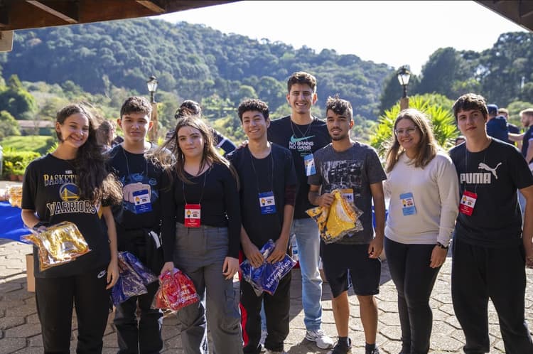 Grupo de oito jovens posa ao ar livre em um evento, usando crachás e roupas casuais, com montanhas e vegetação ao fundo; alguns seguram sacolas e camisetas embaladas.