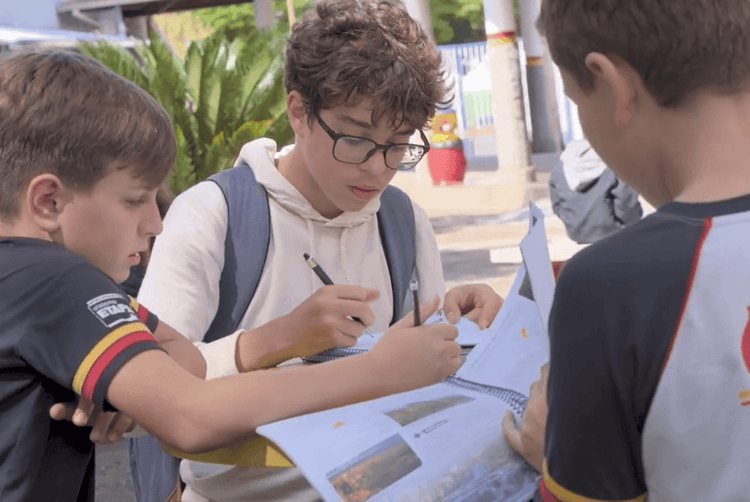 Três estudantes em um ambiente externo analisam uma folha com informações e anotações; o jovem ao centro, de óculos e mochila, escreve enquanto os outros dois observam.