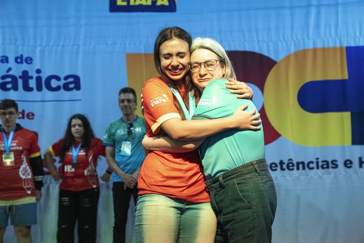 Duas mulheres se abraçam emocionadas em um palco durante um evento, usando camisetas do Sistema Etapa e credenciais no pescoço; ao fundo há outras pessoas e um painel grande com textos e letras coloridas.