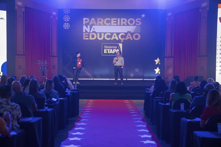 Auditório com público assistindo a uma apresentação; dois homens estão no palco com microfone, e ao fundo há um painel com o texto “Parceiros na Educação” e “Sistema Etapa”, além de um tapete vermelho central com estrelas.