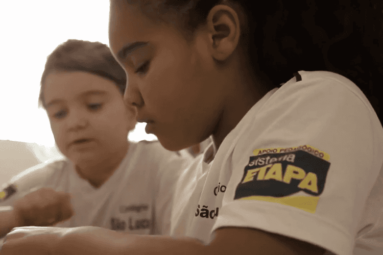 Close em duas crianças estudando juntas; em destaque, uma aluna de perfil usa camiseta branca com o patch “Sistema Etapa” no braço.