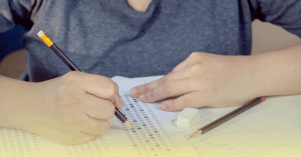 Uma pessoa preenchendo um cartão de respostas de múltipla escolha com um lápis, com uma borracha e outro lápis sobre a mesa.