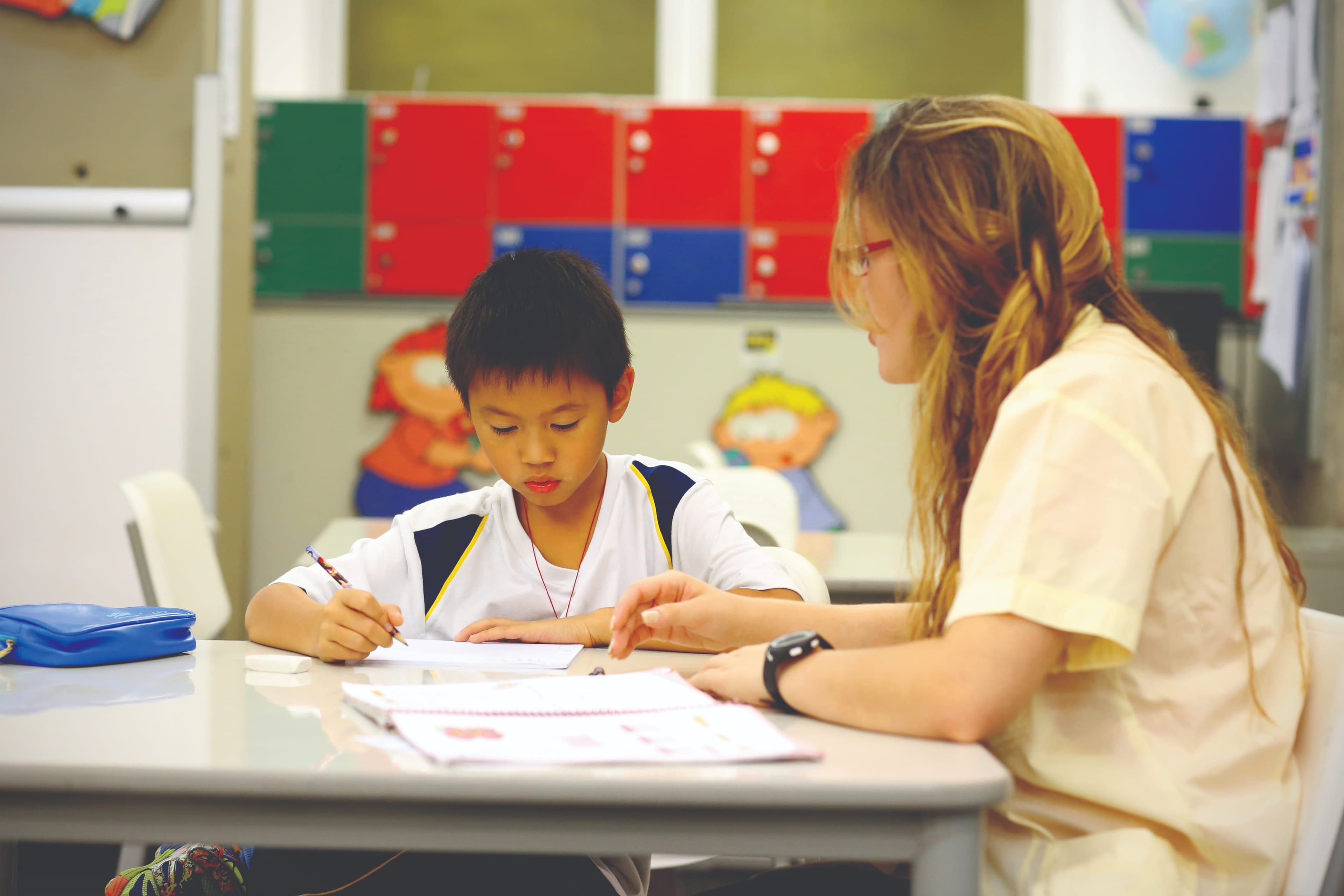 Uma criança escrevendo em uma folha na sala de aula, enquanto uma professora acompanha e orienta ao lado.