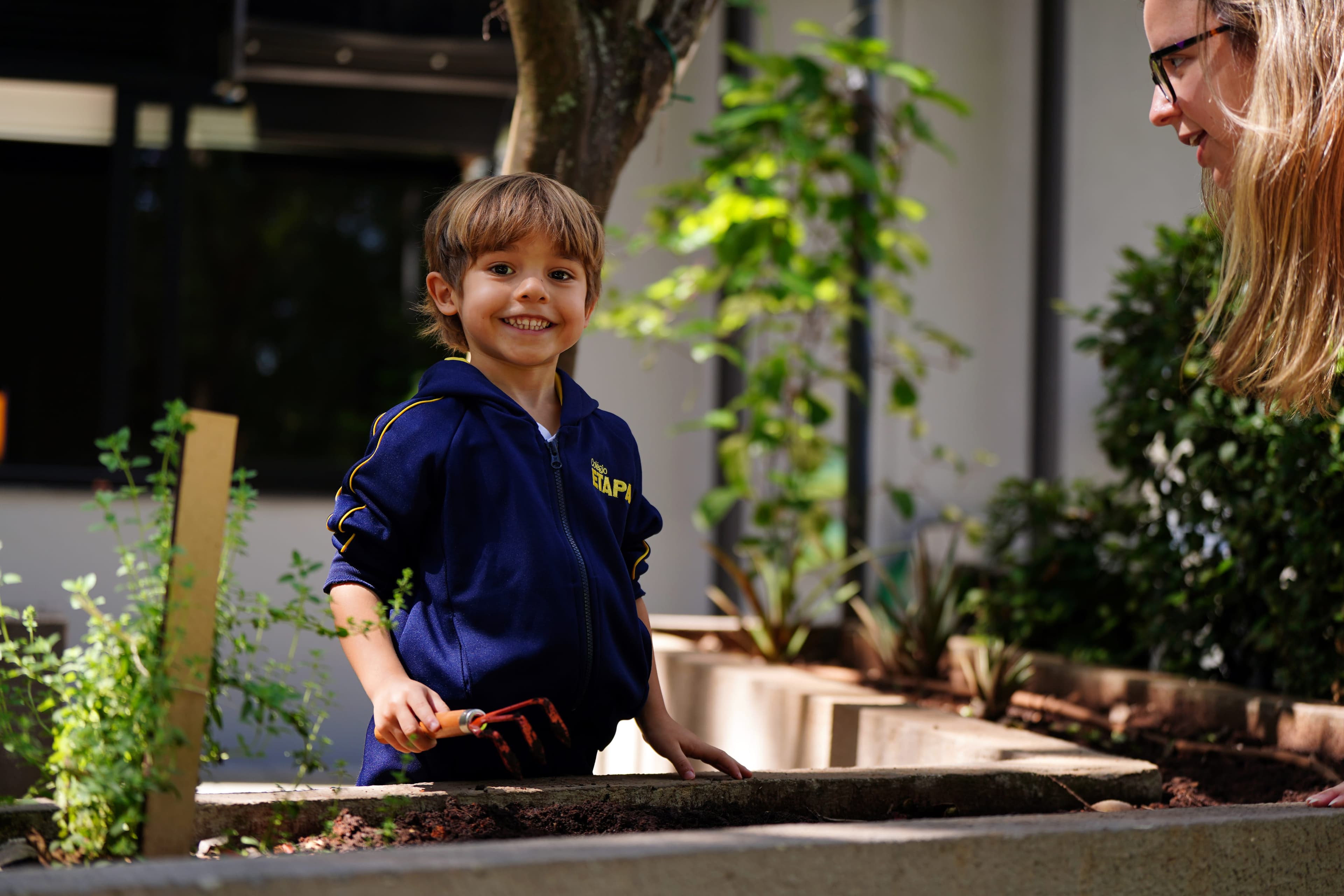 Uma criança sorrindo em um jardim, segurando uma pequena pá, enquanto uma adulta observa ao lado.